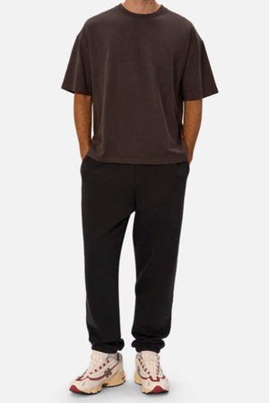 Wearing an oversized dark brown tee, Industrie The New Del Sur Washed Trackpant in black with elastic cuffs, and white sneakers with red and beige accents, a person stands hands-in-pockets against a plain background.