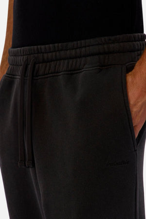 Close-up of a person wearing Industrie The New Del Sur Washed Trackpant—black, oversized fit with drawstring waist, elasticated cuffed ankles, subtle embroidered logo near the pocket—paired with a black shirt and one hand in the pocket.