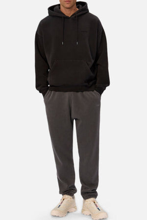 A person stands facing forward in an oversized black hoodie, Industrie The New Del Sur Washed Trackpant in dark gray with logo, and white sneakers, hands in pockets against a plain white background.