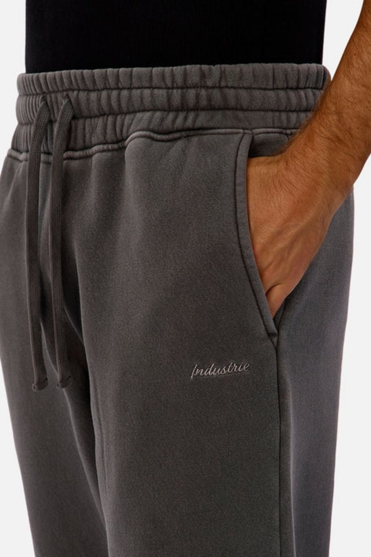 A person wears the Industrie The New Del Sur Washed Trackpant in charcoal grey, featuring an elastic waist with drawstring and “Industrie” embroidery on the thigh, styled with a black top and one hand in the pocket.