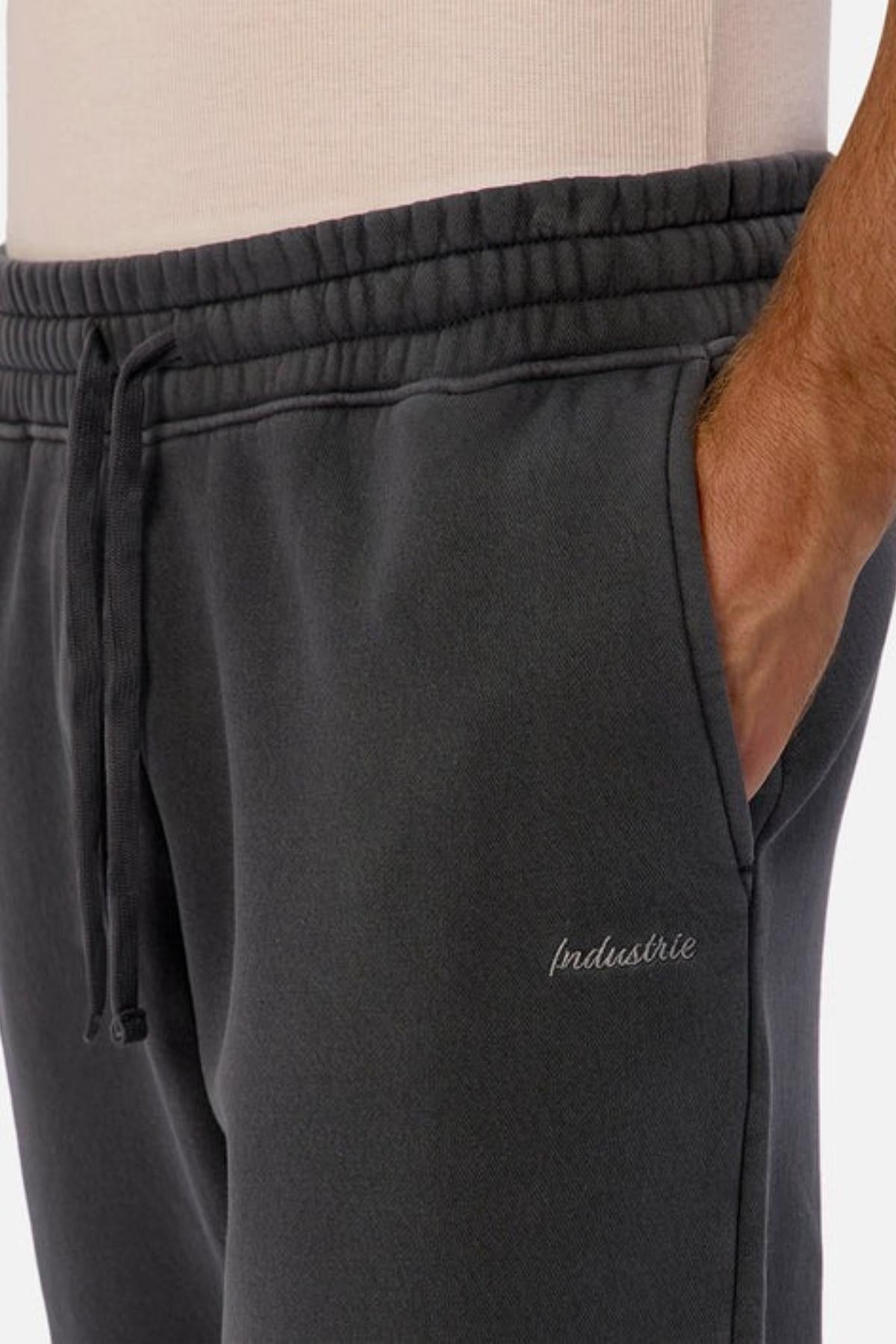 A close-up of a person wearing Industrie The New Del Sur Washed Trackpant featuring an elastic drawstring waist, hand in pocket, "Industrie" stitched on the left leg, and a relaxed oversized fit paired with a light-colored shirt.