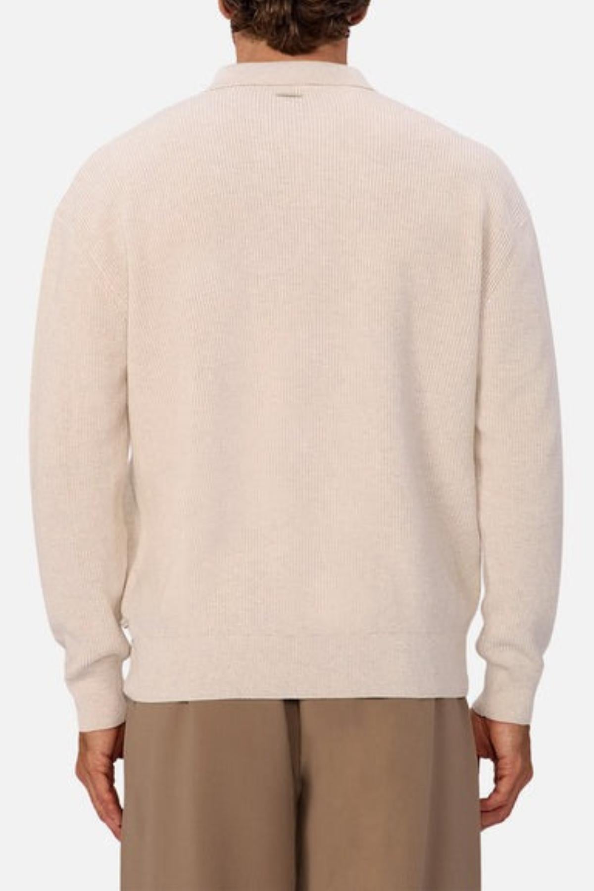 A person is shown from the back wearing the Industrie The New England Polo Knit, a light beige long-sleeve sweater, paired with loose brown pants against a plain light background.