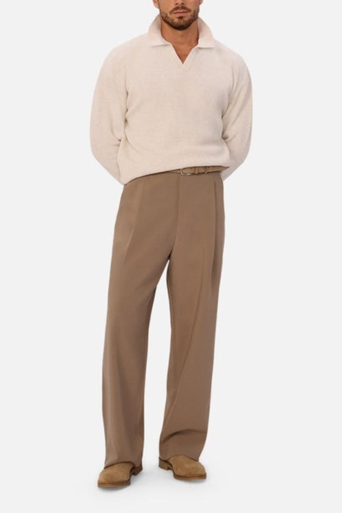 A man stands facing forward in the Industrie The New England Polo Knit, a light beige long-sleeve knit sweater, paired with high-waisted tan belted trousers and light brown shoes. His hands are behind his back against a plain white background.