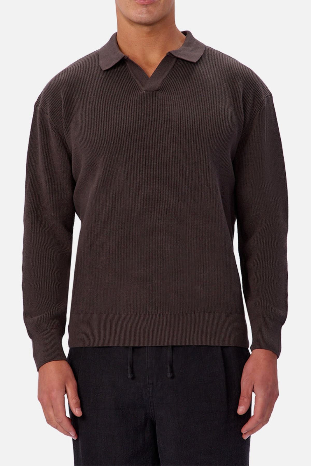Someone wearing the Industrie The New England Polo Knit in dark brown, featuring long sleeves, 100% cotton knit fabric, and a split collar, with black pants, stands against a plain light background.