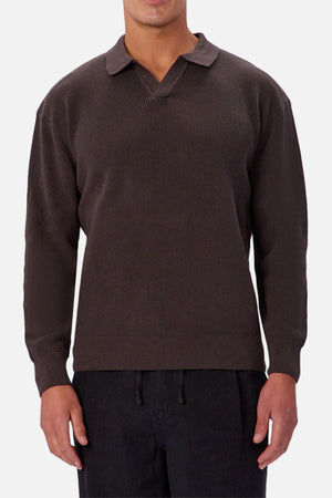 Someone wearing the Industrie The New England Polo Knit in dark brown, featuring long sleeves, 100% cotton knit fabric, and a split collar, with black pants, stands against a plain light background.