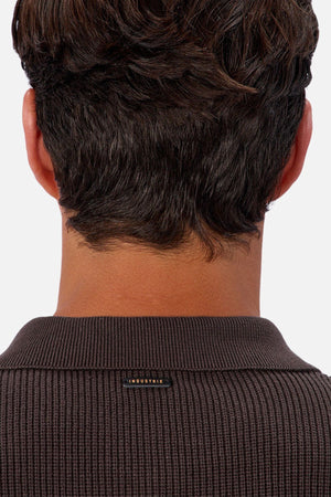A close-up of the back of a person's head and neck shows short dark brown hair and the ribbed brown collar of the Industrie The New England Polo Knit, featuring a small "INDUSTRIE" label. The background is plain and light-colored.
