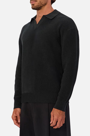 A man stands against a light background in black pants and the Industrie The New England Polo Knit, a black long-sleeve sweater made from 100% cotton knit. His face is partially out of frame.