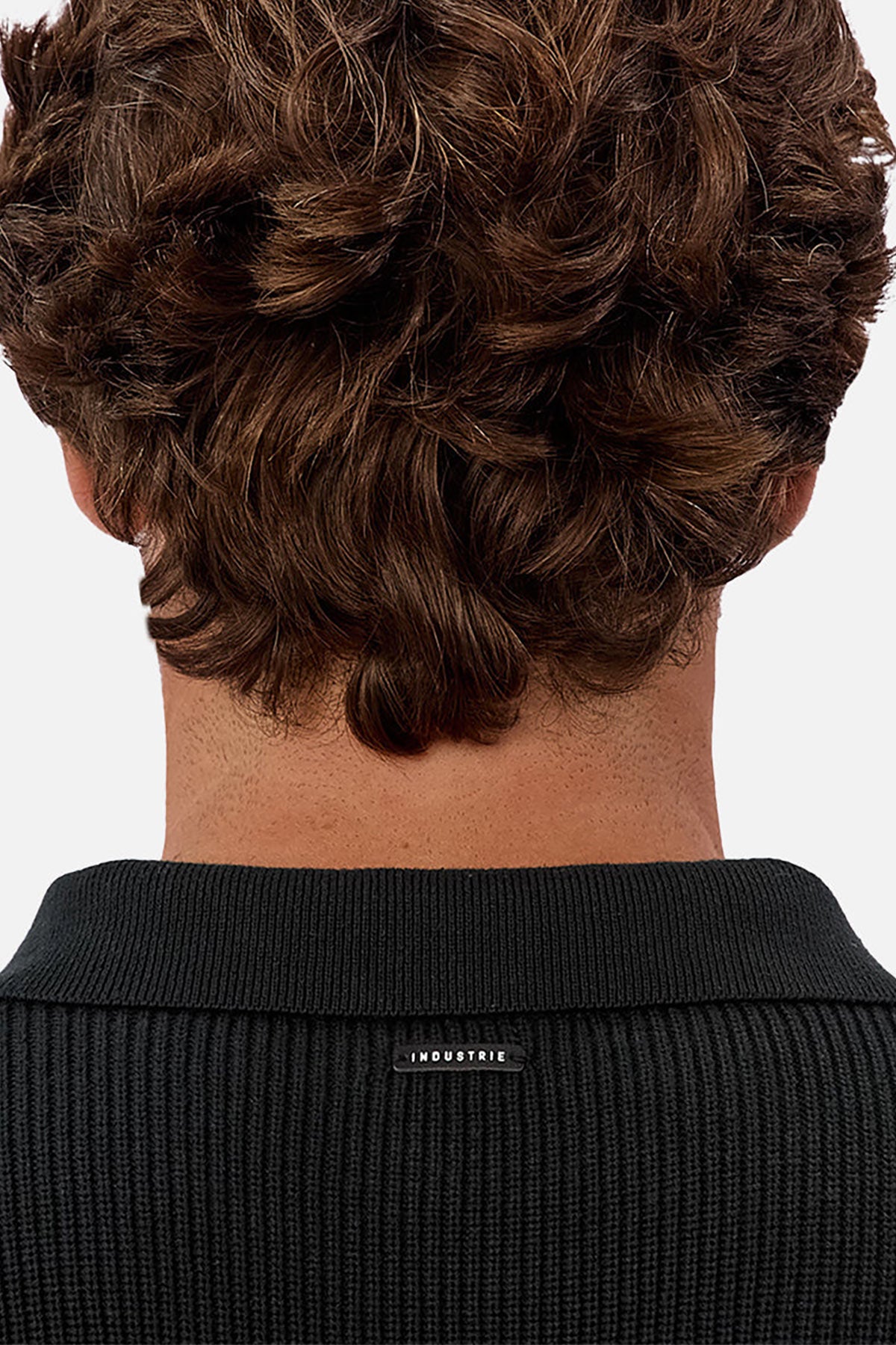 The back view shows a person's head with short, wavy brown hair, wearing the Industrie The New England Polo Knit—a black textured shirt featuring a high-quality knit fabric and a small collar label with the product name.