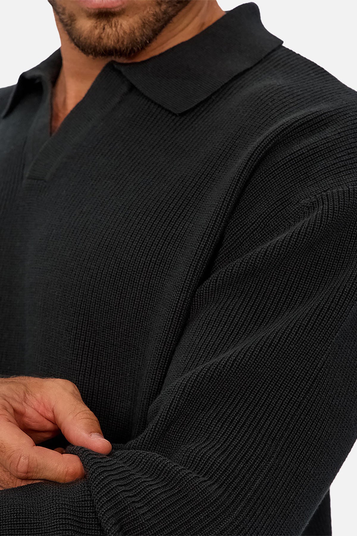 A man wears the Industrie The New England Polo Knit, a textured black long-sleeve collared shirt, lightly pinching its high-quality knit sleeve with his left hand. His face is partially visible against a plain white background.