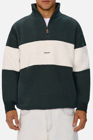 Man wearing the Industrie The New Luton Track Top in dark green and white, "Industrie" on the chest, paired with light pants. This relaxed fit quarter zip top features a partially zipped front and hands resting casually at his sides.