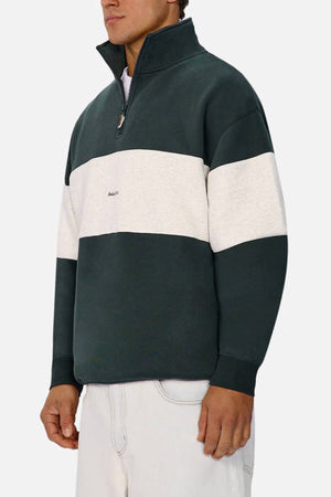 Someone wears the Industrie The New Luton Track Top in dark green and white, featuring a relaxed fit and small chest logo, paired with light-colored pants. The quarter zip design leaves the face partially out of frame.