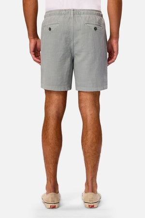 A person stands facing away, wearing Industrie The New Mallorca Short—lightweight, breathable men’s cotton shorts in light gray featuring two back pockets with button closures—paired with beige slip-on shoes and a white shirt. The background is plain white.