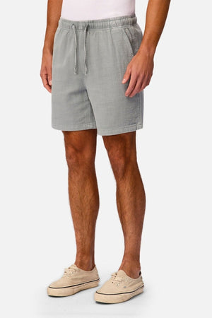 A person wearing Industrie The New Mallorca Short—a pair of lightweight, breathable men’s cotton shorts in light gray—paired with a white shirt and beige slip-on shoes stands against a plain white background.