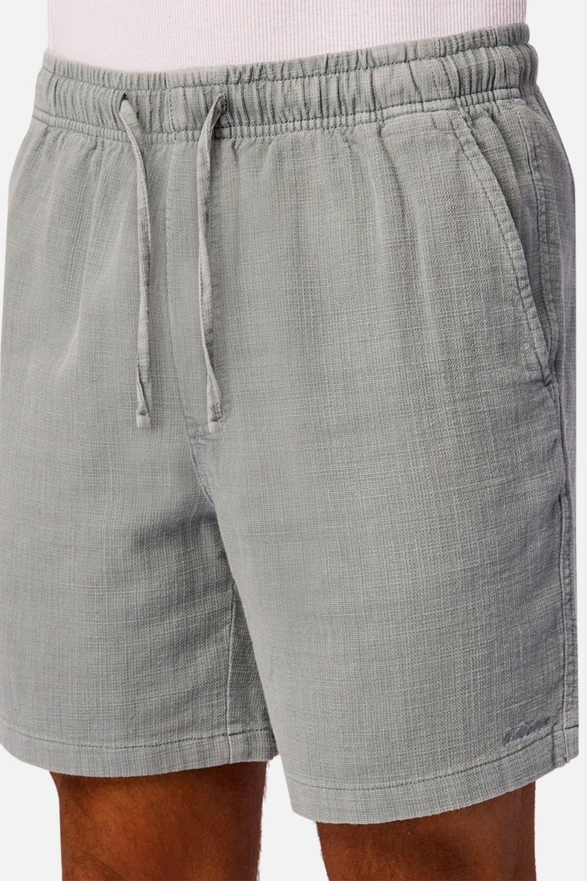 A person is shown wearing Industrie The New Mallorca Short in light gray, featuring a textured design, elastic waistband with drawstring, and practical side pockets, paired with a white shirt. The lightweight and breathable shorts provide both comfort and style. Only the lower torso and upper legs are visible.