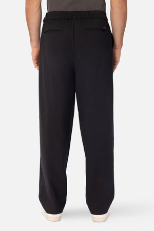 A person is seen from behind wearing Industrie The New Milano Pant—loose black pants with an elastic waistband and two welt pockets. White shoes finish this professional look against a plain light background.