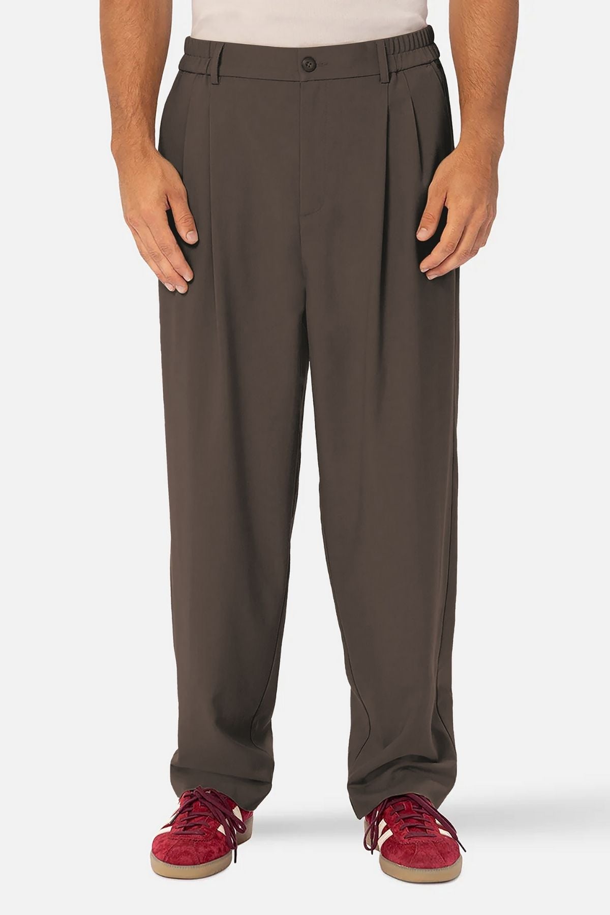 A person wears loose-fitting Industrie The New Milano Pant in brown, paired with a white shirt and red sneakers, standing against a plain white background. The image is cropped from the shoulders down.