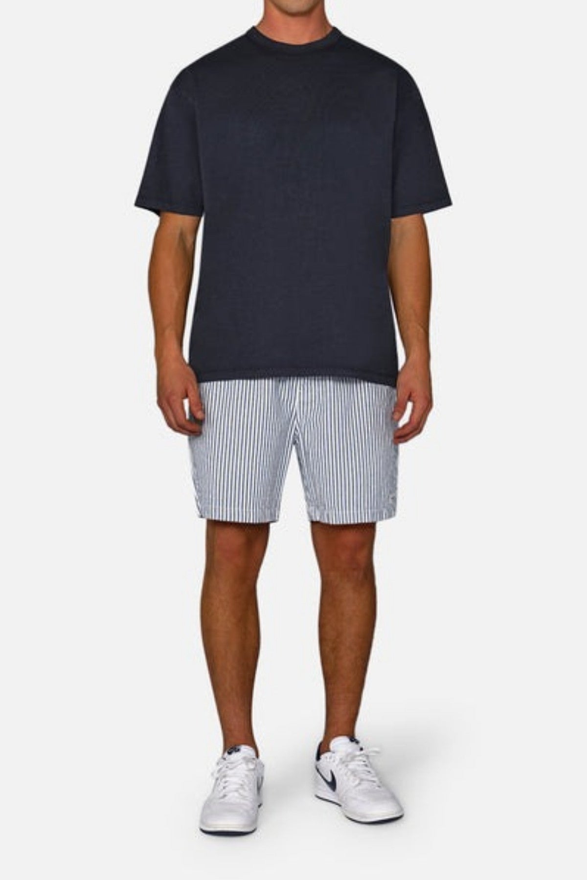 A person stands against a plain background wearing a black T-shirt and the Industrie The New Southwest Short, which features white and blue vertical stripes along with an elasticated waistband. Paired with white sneakers, the outfit exudes a casual and relaxed vibe.