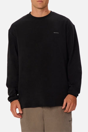 A person stands against a white background wearing an oversized fit black Industrie The Newport Long Sleeve Tee with small white chest text and light brown pants.