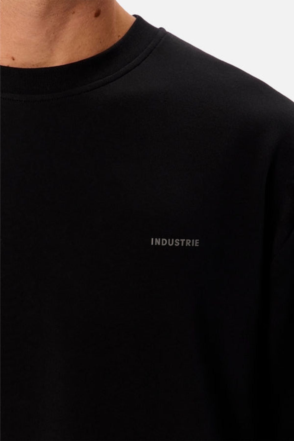 Close-up of a person wearing the Industrie The Newport Long Sleeve Tee, an oversized black crewneck with "INDUSTRIE" printed in small gray letters on the left chest. Only the lower neck and upper chest are visible.
