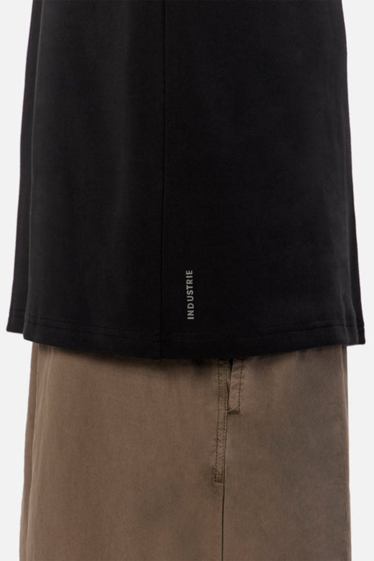 A close-up shows only the lower torso of a person wearing the Industrie The Newport Long Sleeve Tee paired with taupe pants. The tee features a small vertical "INDUSTRIE" logo on its hem, highlighting its oversized fit and heavyweight finish.