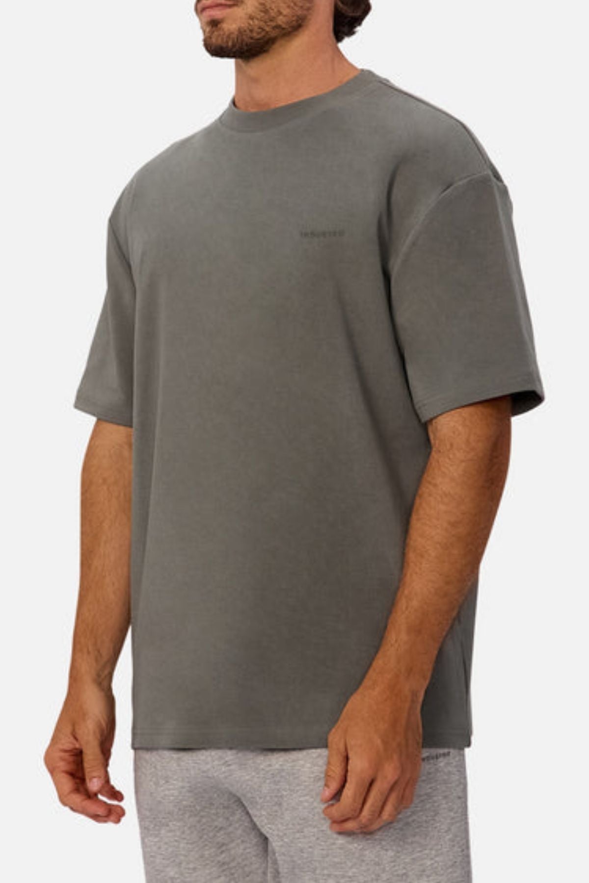 A man wears The Newport Tee by Industrie in a loose, dark gray design with short sleeves and light gray shorts, standing against a plain white background. Its oversized fit and heavyweight fabric provide a relaxed yet structured look.