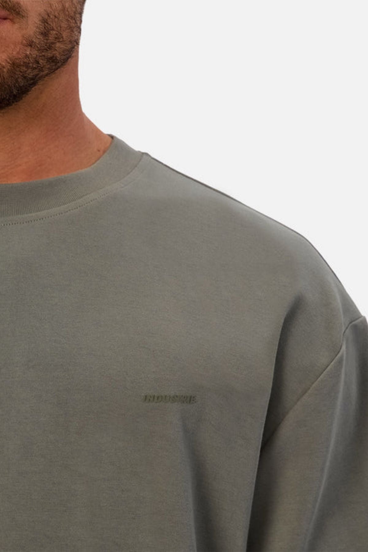A close-up of a person wearing the Industrie The Newport Tee in light grey-green heavyweight fabric, featuring subtle embossed chest text, partially visible against a plain white background.