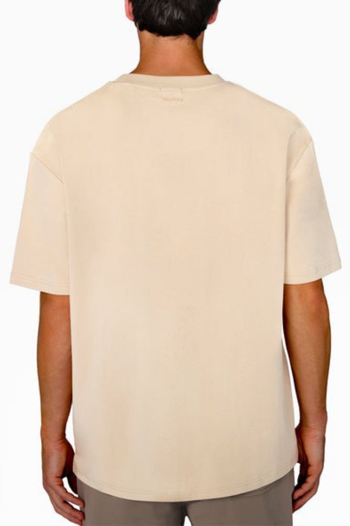 Rear view of a person wearing the Industrie The Newport Tee in a plain beige color, paired with brown pants, standing against a white background. This T-shirt, made from 100% cotton, features an oversized fit with short sleeves. The person's arms hang relaxed at their sides, showcasing an effortless casual comfort and style.