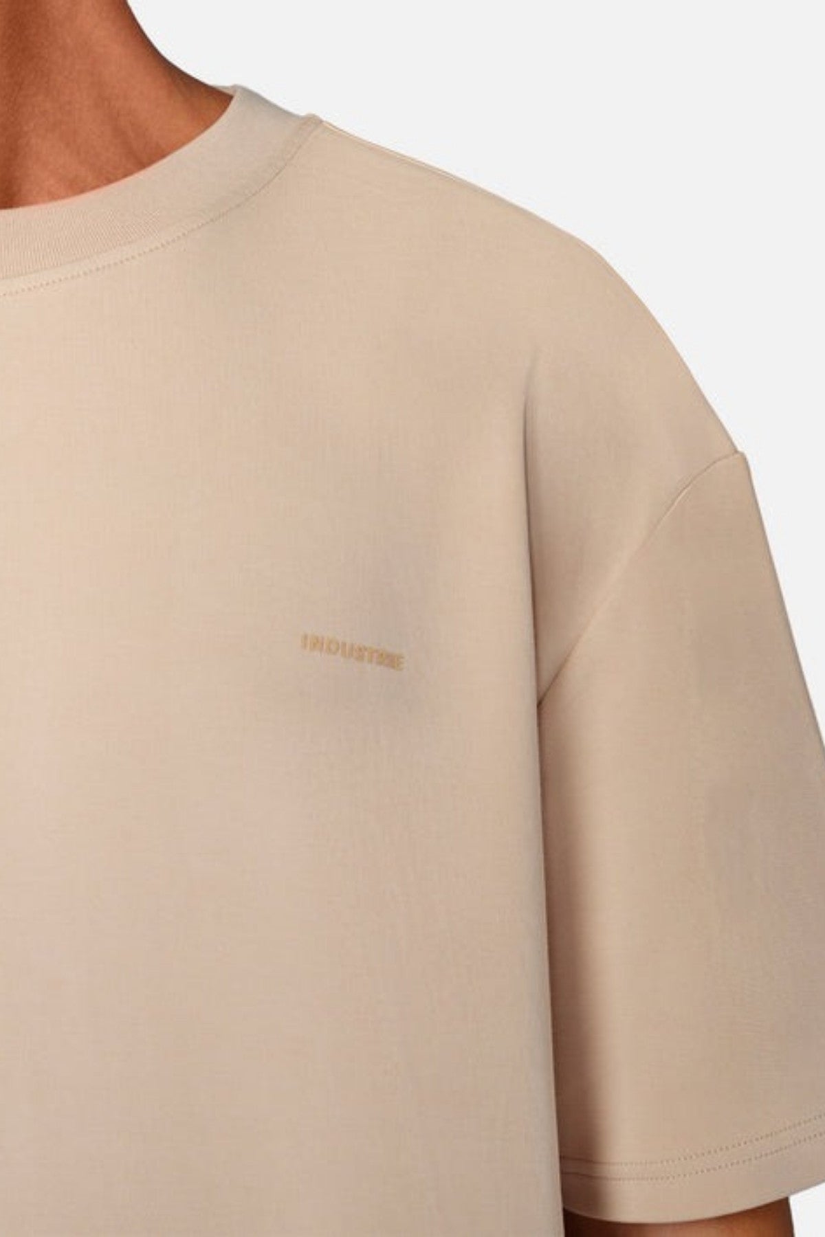 Close-up of a person wearing the Industrie The Newport Tee, a beige T-shirt featuring an oversized fit and a subtle branded logo on the chest. The fabric appears soft, crafted from 100% cotton, with short sleeves that highlight its minimalist design.