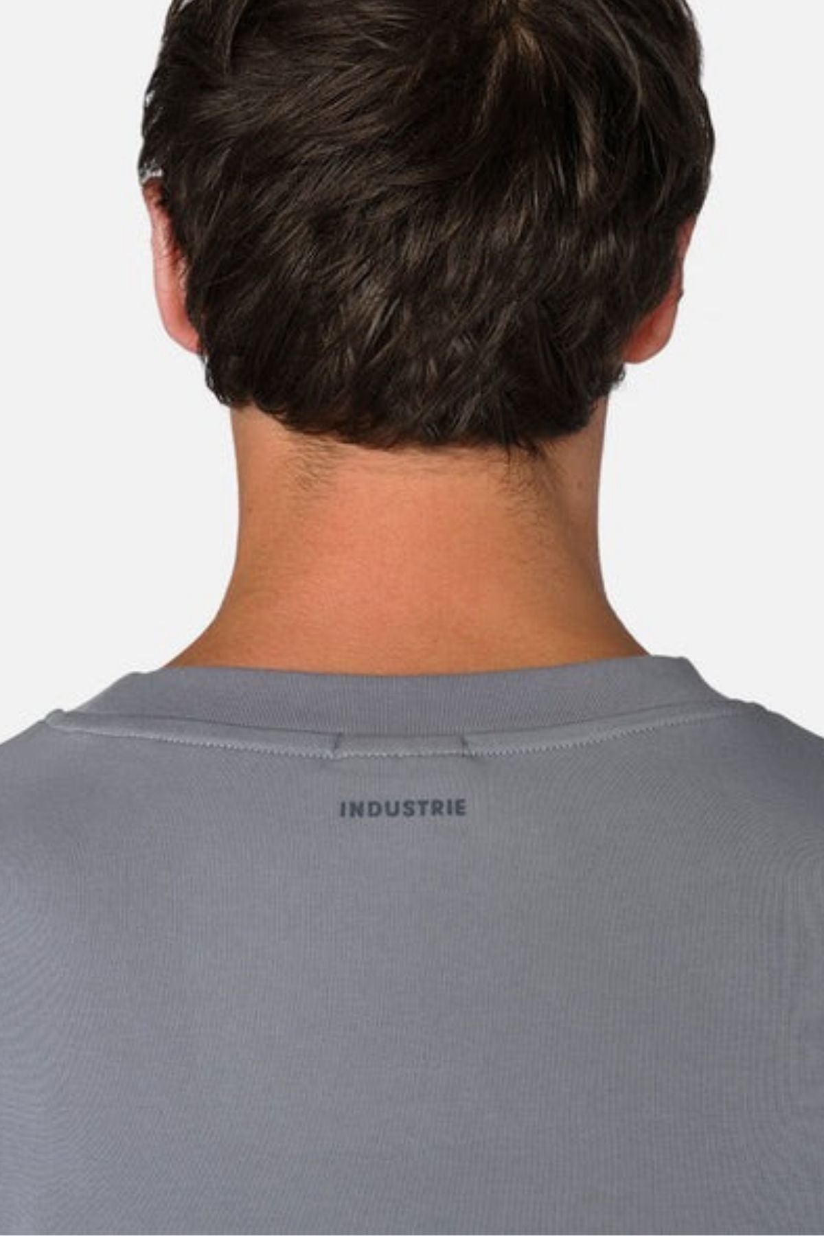 The image features the rear view of an individual wearing a gray oversized tee, identified as "Industrie The Newport Tee," with "INDUSTRIE" printed near the neckline. The 100% cotton shirt accentuates their dark hair against a simple backdrop.