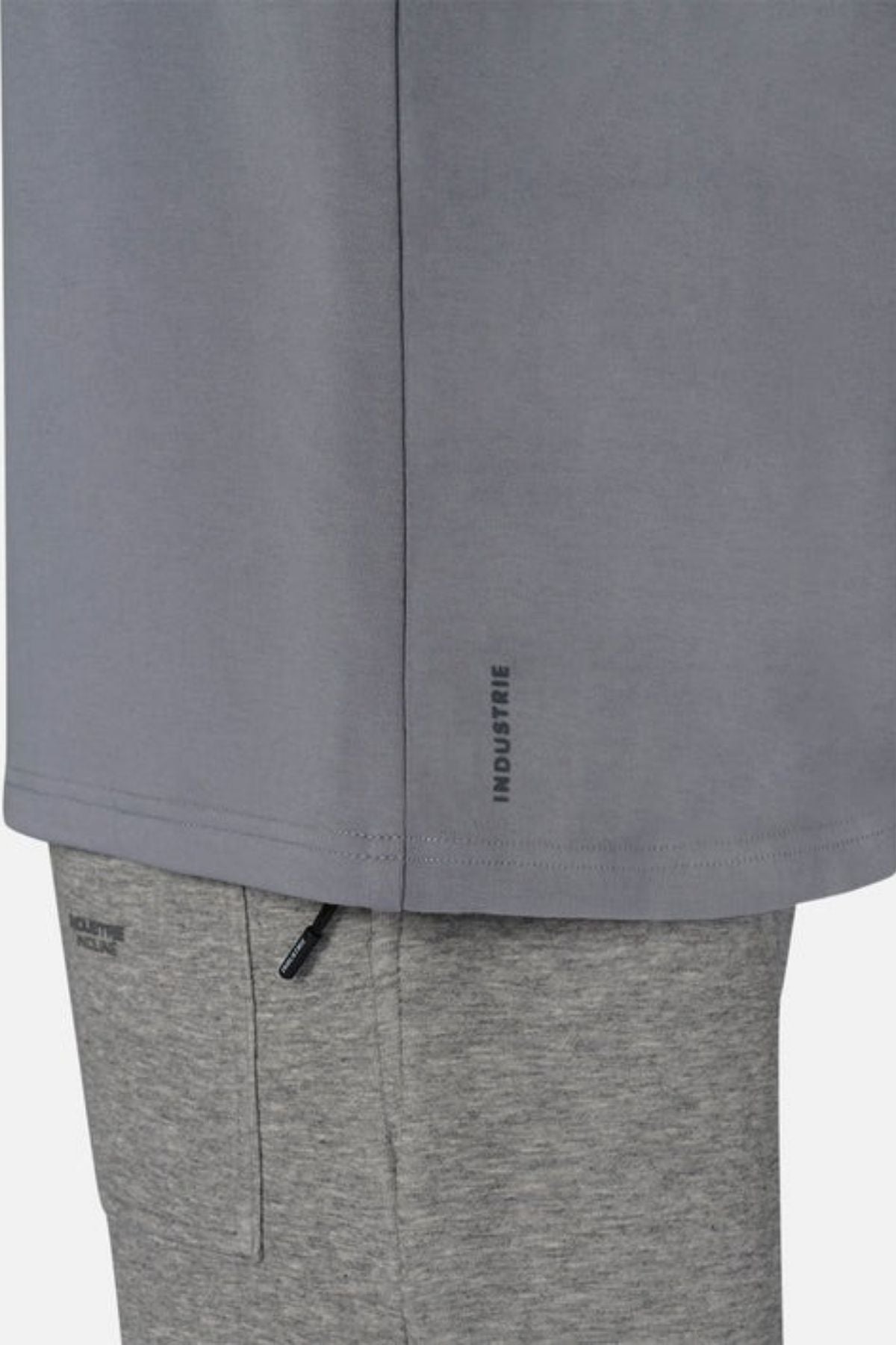 A close-up captures a person donning an oversized Industrie The Newport Tee in gray, paired with lighter gray pants. The shirt, made entirely from cotton, showcases "INDUSTRIE" near the hemline. The pants include a pocket accented by a black zipper. The backdrop is simple and light-hued.