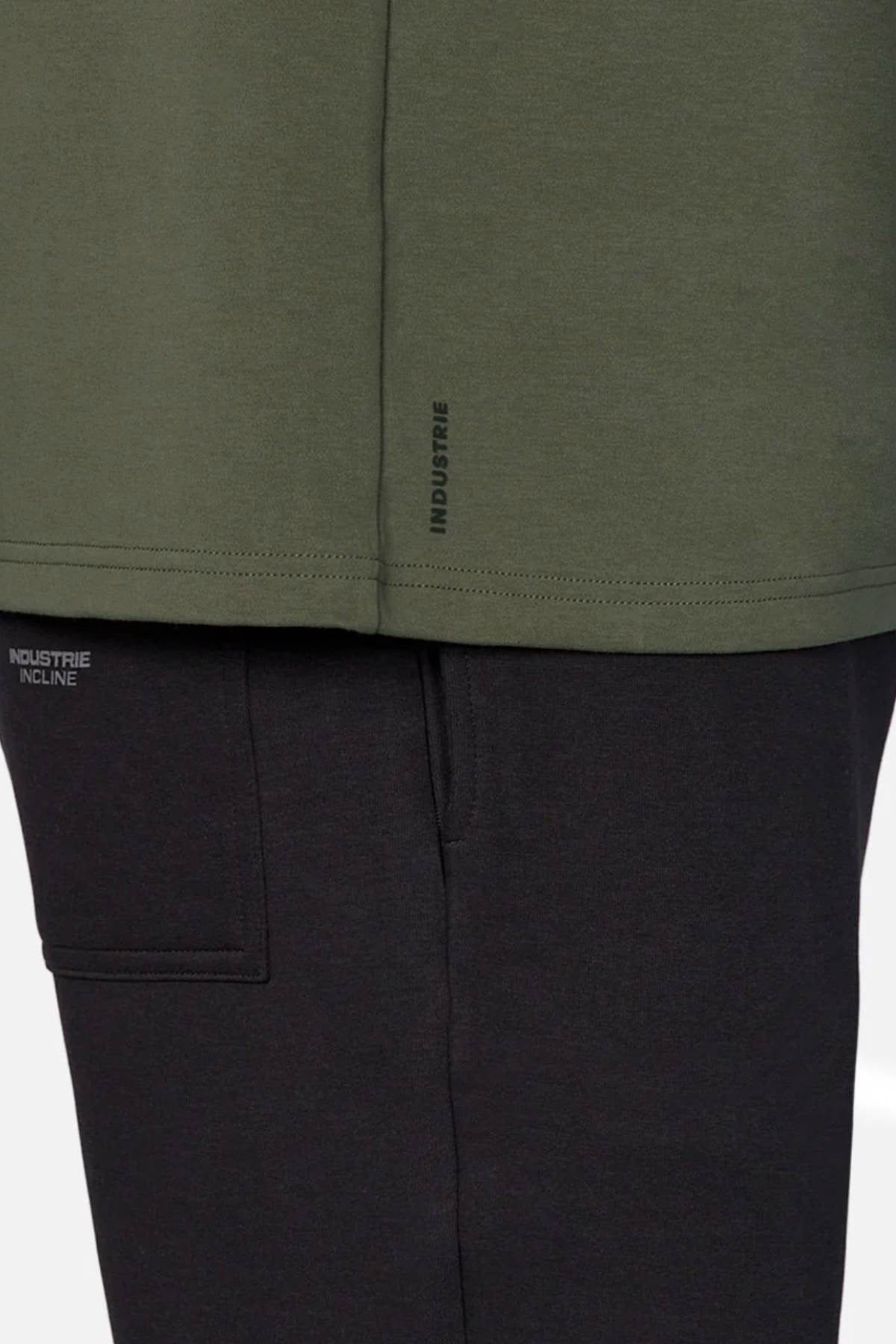 A close-up of a person wearing an oversized green Industrie The Newport Tee and black pants, with "INDUSTRIE" and "INDUSTRIE INCLINE" in small print on the 100% cotton tee’s hem and the pants pocket.