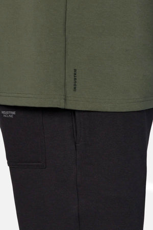 A close-up of a person wearing an oversized green Industrie The Newport Tee and black pants, with "INDUSTRIE" and "INDUSTRIE INCLINE" in small print on the 100% cotton tee’s hem and the pants pocket.