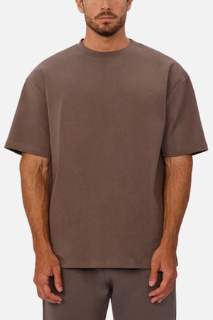 A man wearing an oversized fit, short-sleeve, brown Industrie The Newport Tee with matching brown pants stands against a plain white background.