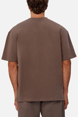A person with short curly hair is shown from behind, wearing an oversized Industrie The Newport Tee and matching brown shorts against a plain, light background.