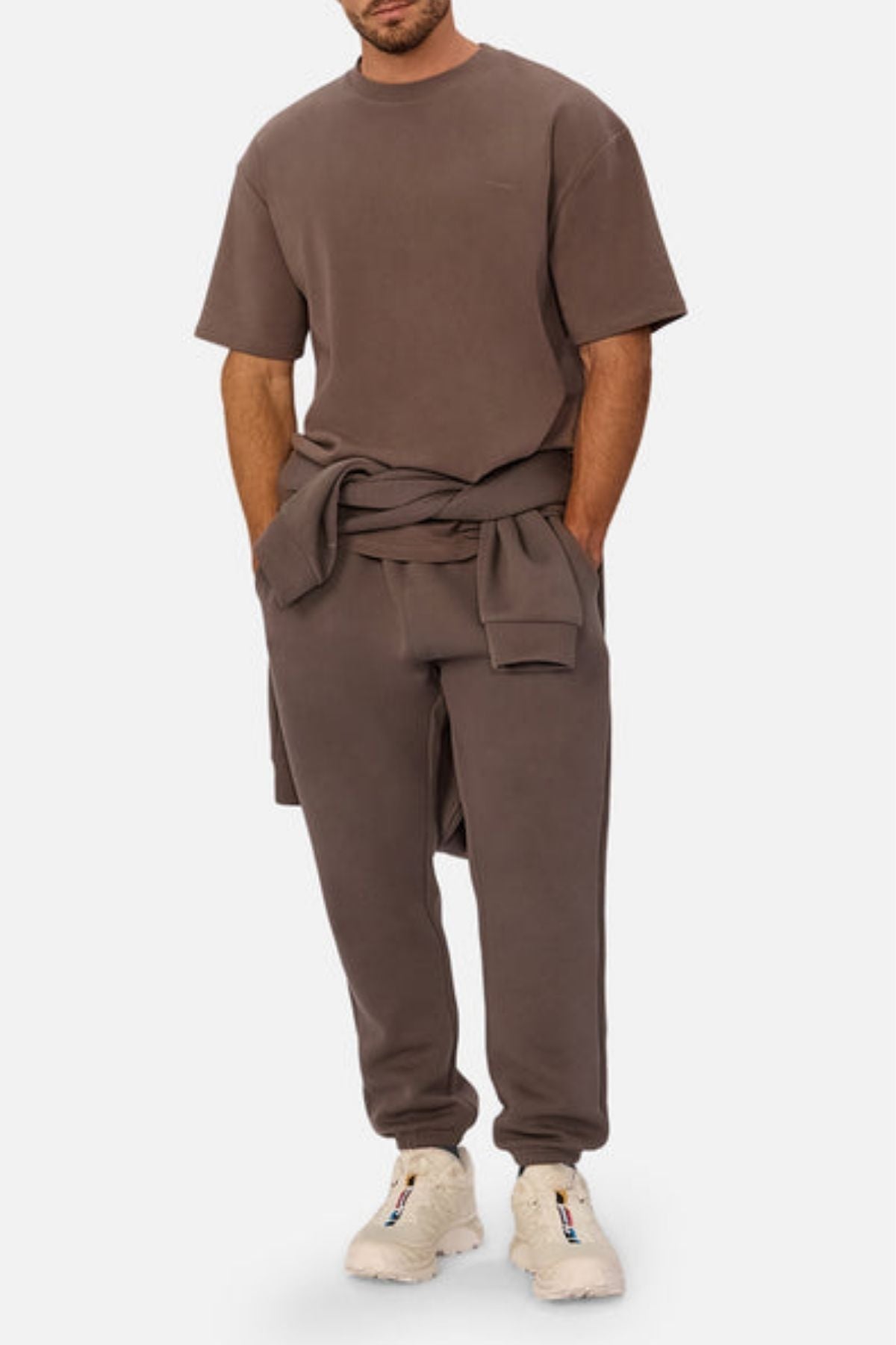 A man wearing the Industrie The Newport Tee, an oversized 100% cotton brown t-shirt, and matching sweatpants with a sweatshirt tied at his waist and white sneakers stands against a plain white background.