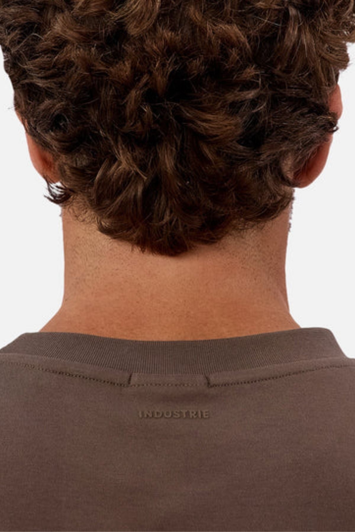 The image shows the back of a person with short, curly brown hair wearing the Industrie The Newport Tee—an oversized brown 100% cotton shirt with "INDUSTRIE" printed near the upper back collar.