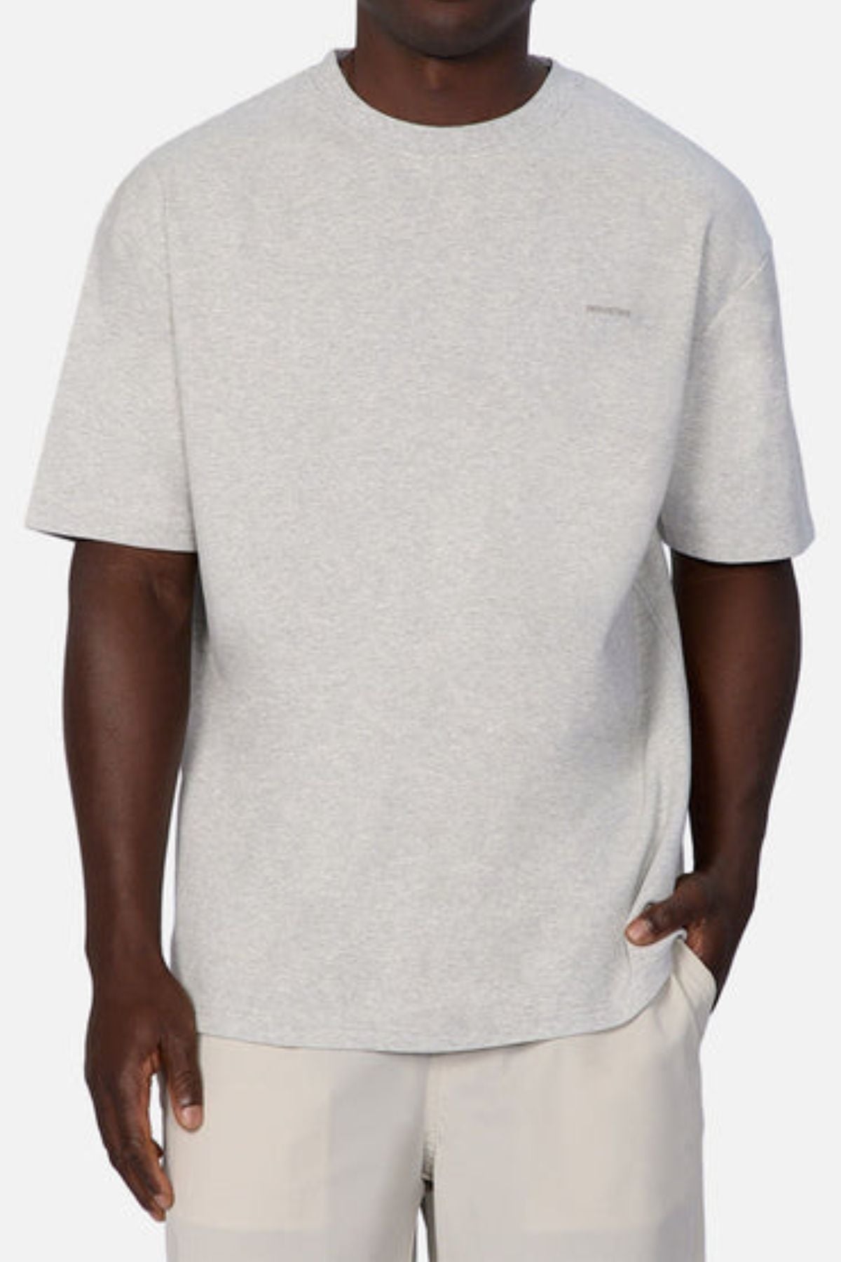 A person dressed in an Industrie The Newport Tee, known for its oversized fit, paired with beige pants, stands against a plain background. The shirt is light gray and crafted from 100% cotton, showcasing a discreet logo on the left chest. Their left hand is tucked into their pants pocket.