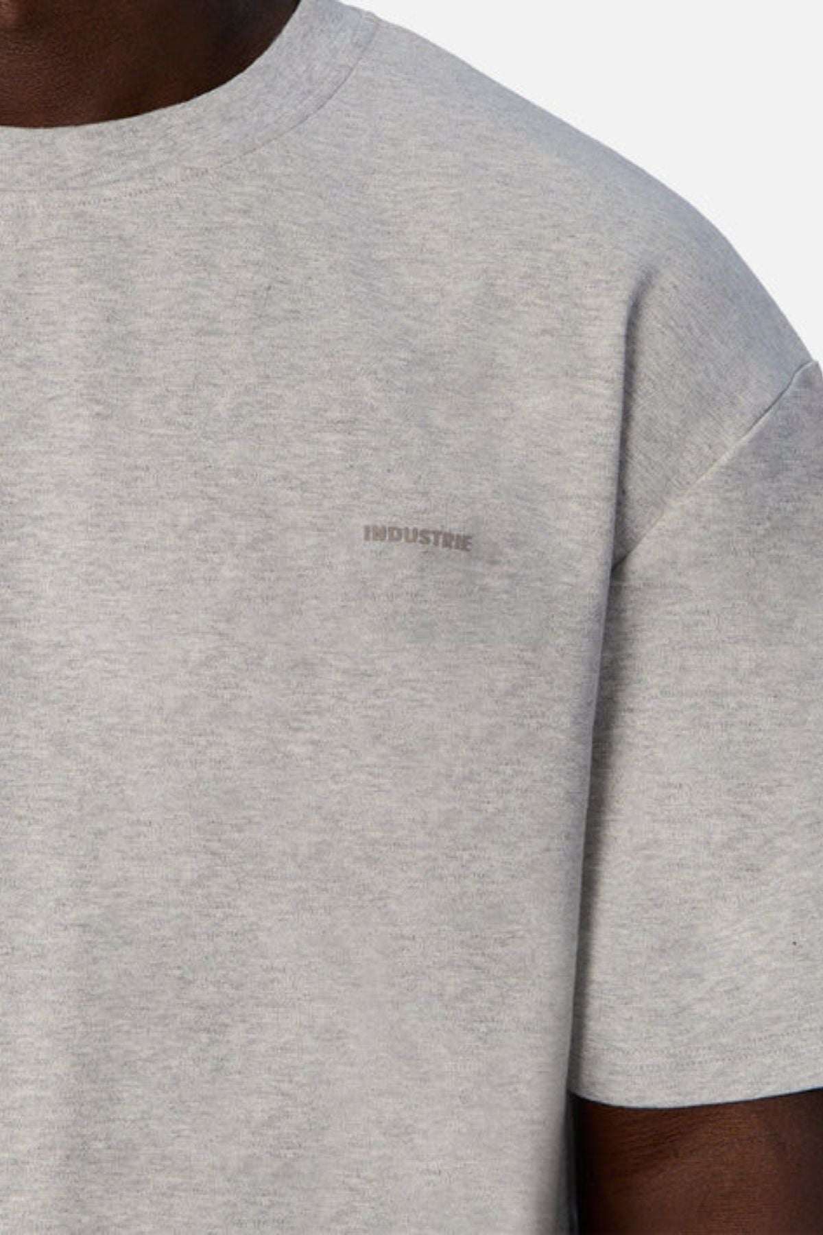 Close-up of a person wearing "Industrie The Newport Tee" in light gray, featuring the word "INDUSTRIE" subtly printed on the chest. Made from 100% cotton, the top half of the T-shirt and part of the sleeve reveal its oversized fit against a plain background.