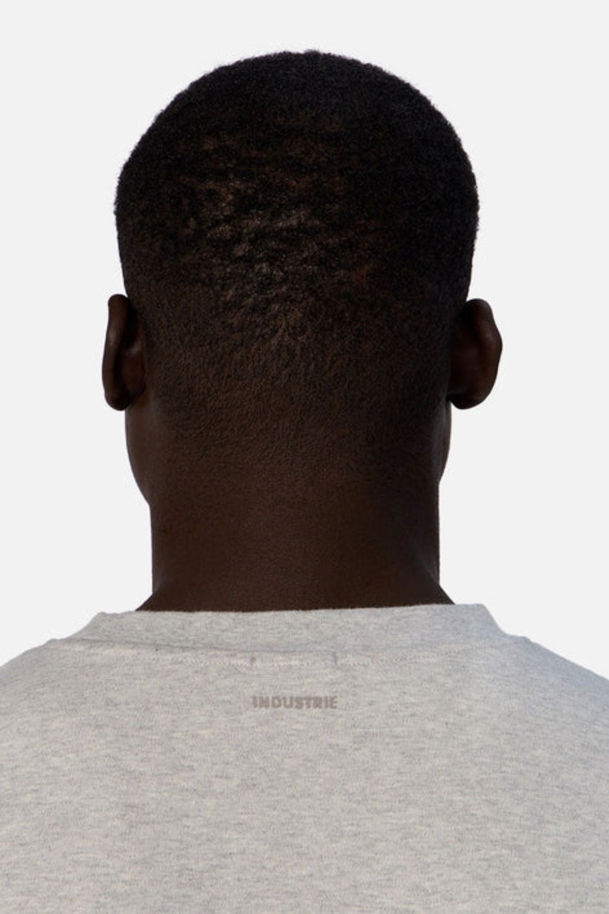 A person with short, dark hair is depicted from behind, dressed in an oversized light gray Industrie The Newport Tee that features "INDUSTRIE" printed on the back. This t-shirt is made entirely of cotton and set against a plain white background.