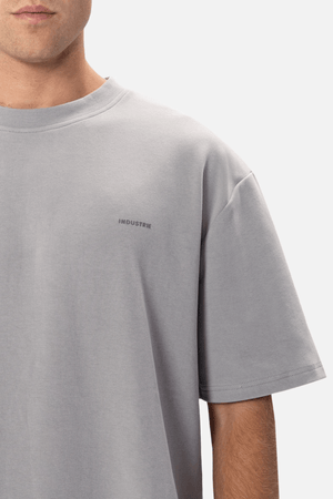 A person is donning Industrie The Newport Tee, an oversized fit, light grey T-shirt with short sleeves. Made from 100% cotton, it features "INDUSTRIE" printed in small black letters on the left chest. The background is plain white, showcasing only the upper torso and part of the face.