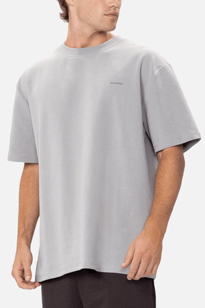 A person with short hair is wearing a loose, light gray T-shirt and black shorts. The T-shirt is an Industrie The Newport Tee, made from 100% cotton, featuring a small, subtle logo on the chest. The person is standing and looking to their right against a plain, light-colored background.