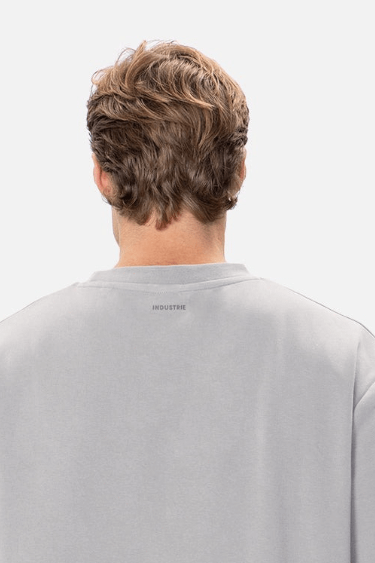 A person with wavy brown hair is seen from behind, wearing an oversized light gray sweatshirt. The word "INDUSTRIE" is printed in small letters on the back below the neckline. This 100% cotton piece resembles Industrie The Newport Tee and stands out against the plain white background.