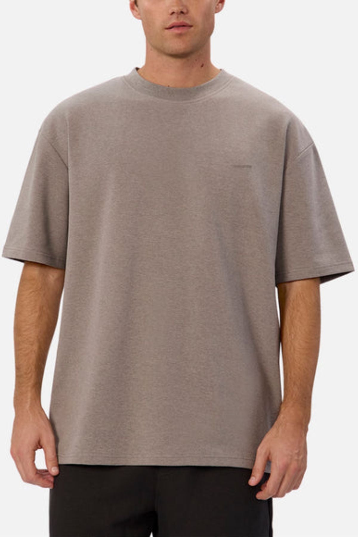 A person is wearing Industrie The Newport Tee, showcasing an oversized fit in plain beige, made from 100% cotton. This t-shirt includes a subtle chest logo and is paired with black pants against a white background.