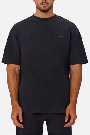 A man wears an oversized, short-sleeve black Industrie The Newport Tee made from heavyweight fabric with a subtle chest logo, paired with black pants, standing against a plain light background.
