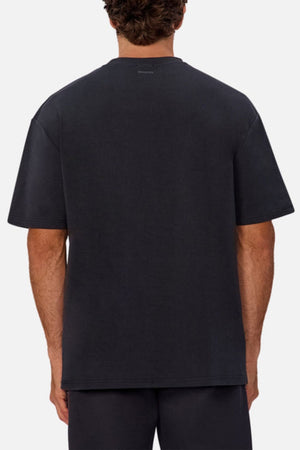 A person with curly brown hair is shown from the back, wearing an oversized fit, short-sleeve Industrie The Newport Tee in black and black shorts against a plain white background.