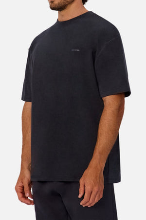 A man wears an oversized, short-sleeved black Industrie The Newport Tee with subtle chest text, paired with black pants, against a plain light background.