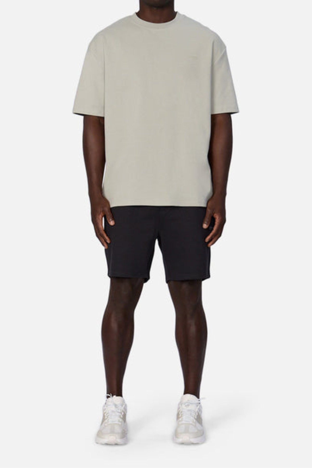 A person stands against a plain backdrop in Industrie's The Newport Tee in light gray, black shorts, and white sneakers. The oversized fit and heavyweight fabric of the tee enhance the casual style. Their arms rest relaxed at their sides.