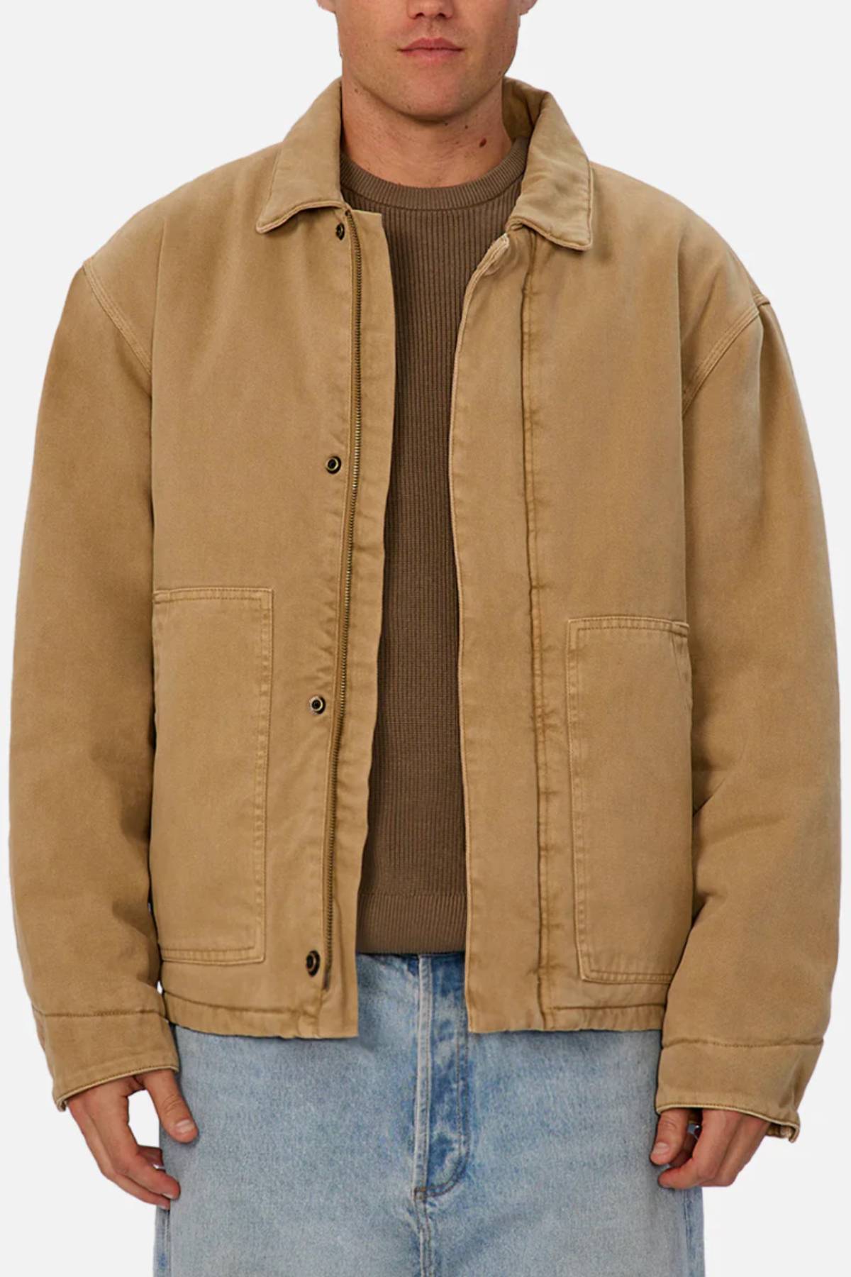A person wears the Industrie The Oklahoma Jacket in tan, featuring large front pockets and a collar, over a brown ribbed sweater and light blue jeans against a plain background for an effortlessly stylish, casual look.