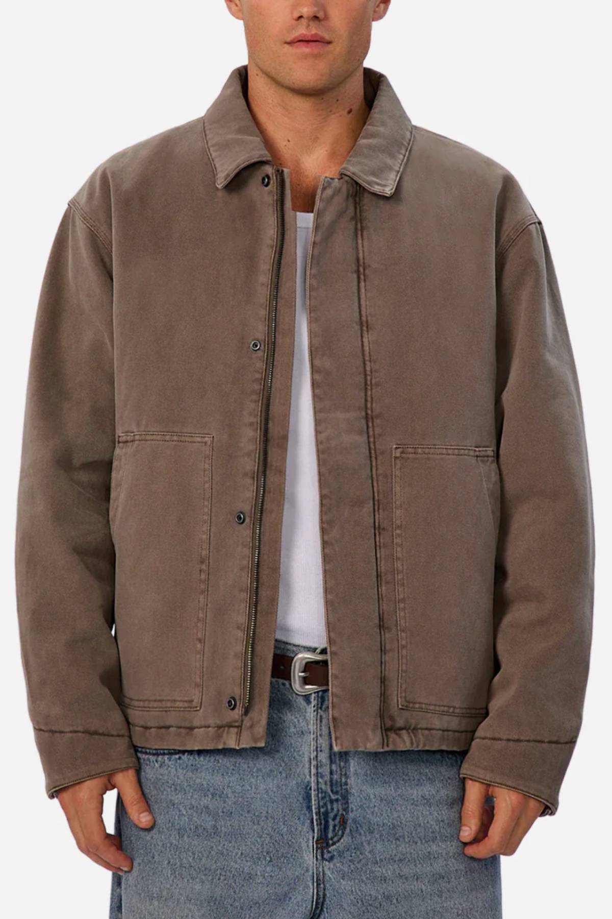 Someone stands facing forward against a plain light background, wearing Industrie The Oklahoma Jacket—a loose-fitting brown jacket with a collar, button closure, and two front pockets—layered over a white shirt and blue jeans.