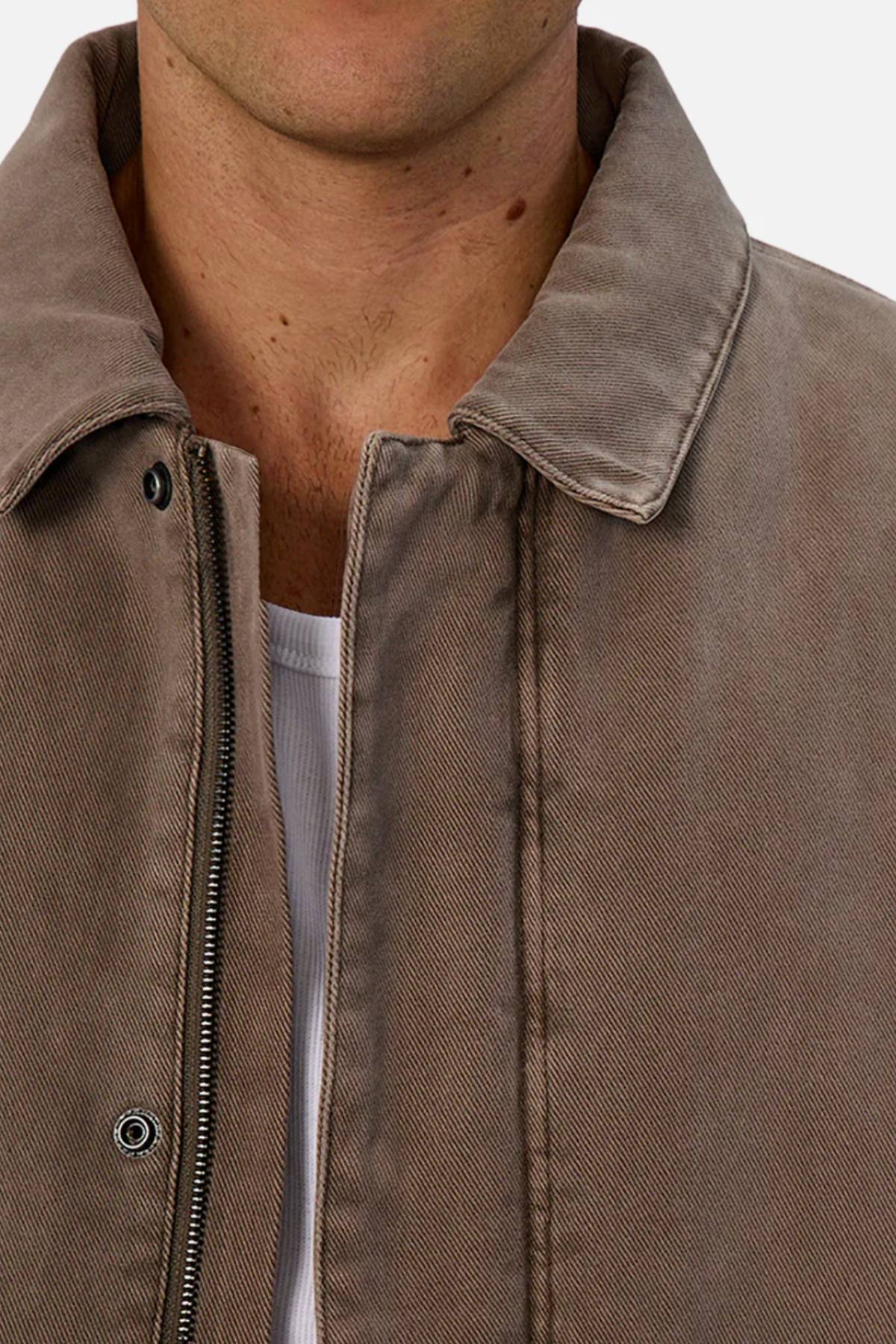 Close-up of a person wearing the Industrie The Oklahoma Jacket—a light brown, casual-fit piece with a zipper and snap buttons over a white ribbed shirt. Only the lower half of the face and upper torso are shown, emphasizing this versatile outerwear.